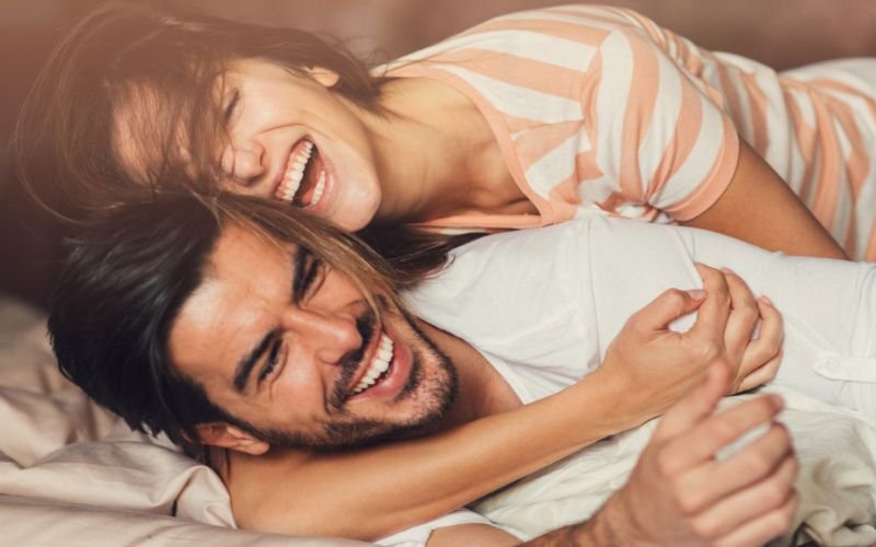 A cheerful couple laughing and cuddling playfully on a bed, showing happiness and intimacy.