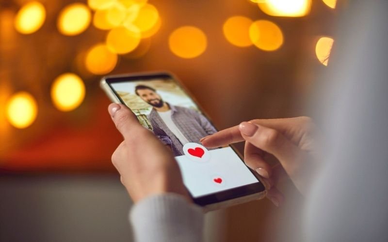 A close-up of a woman using a dating app on her smartphone, tapping the heart icon on a profile with blurred golden bokeh lights in the background.